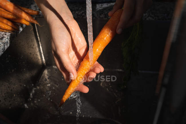 Abgeschnittenes Bild einer Frau, die zu Hause reife Karotten unter sauberem Wasser über der Spüle wäscht — Stockfoto