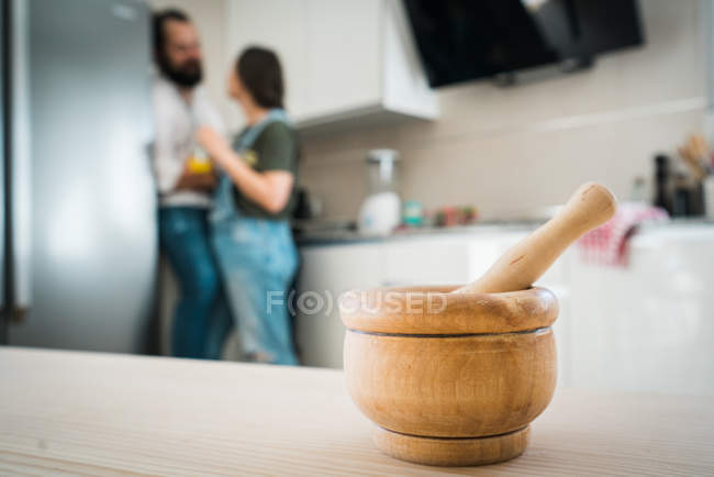 Holzmörtel und Stößel auf Bauholz-Tischplatte auf verschwommenem Hintergrund von Küche und Paar zu Hause platziert — Stockfoto