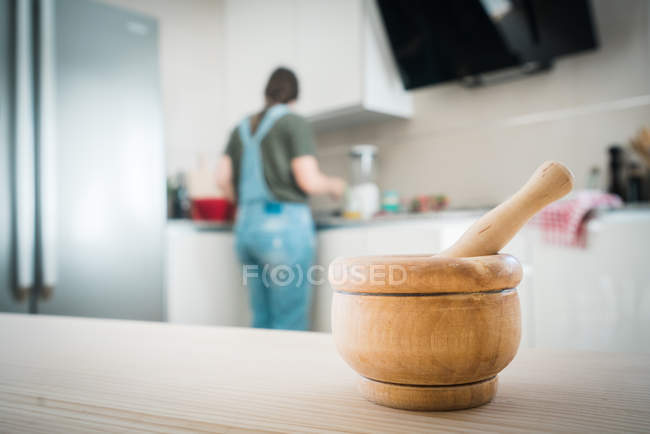 Mortero y mortero de madera colocado sobre una mesa de madera sobre un fondo borroso de la cocina con una mujer detrás de ella en casa - foto de stock