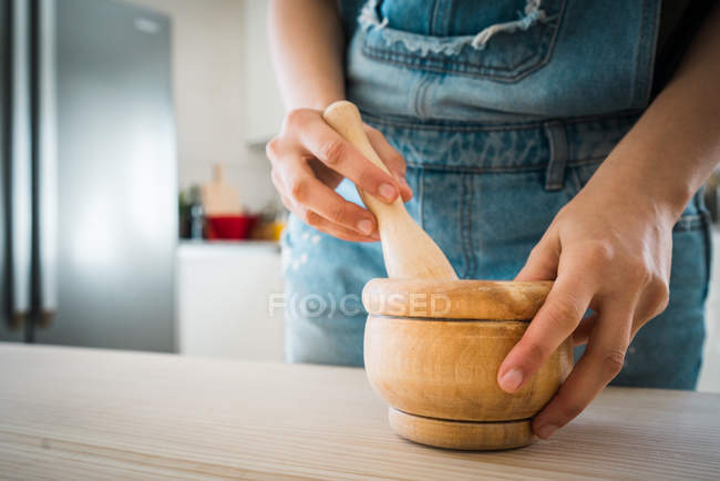 Imagem cortada da mulher usando argamassa de madeira e pilão colocado na mesa de madeira na cozinha em casa — Fotografia de Stock