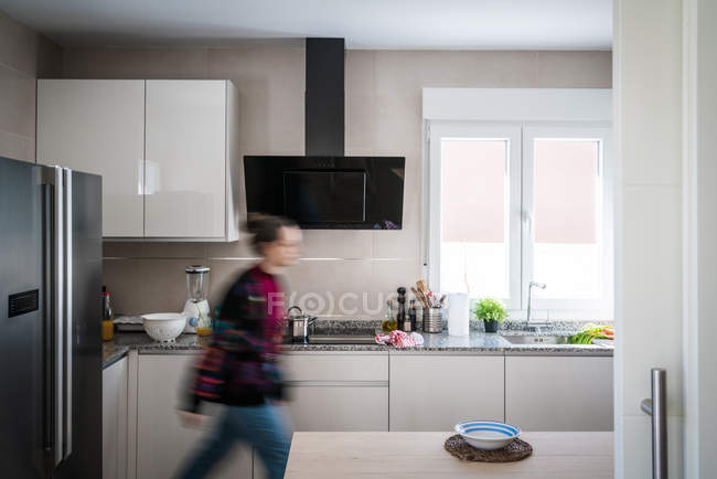 Невпізнавана жінка ходить в інтер'єрі легкої просторої кухні, обставленої білими шафами і різноманітним сучасним обладнанням — стокове фото