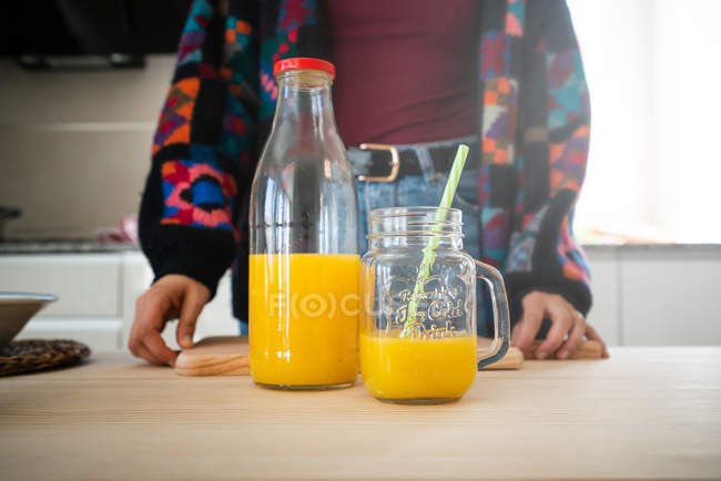 Frasco de suco de laranja fresco gostoso e vidro com palha colocada na mesa de madeira com mulher cortada atrás — Fotografia de Stock