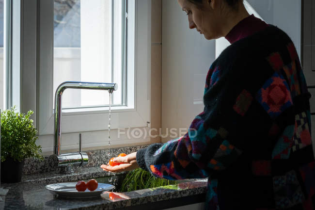 Женщина в разноцветной куртке моет свежие помидоры под чистой водой над раковиной на кухне дома — стоковое фото
