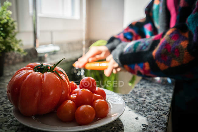Imagen recortada de la mujer en chaqueta colorida lavado de tomates frescos y zanahoria bajo agua limpia sobre el fregadero en la cocina en casa - foto de stock