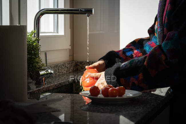 Immagine ritagliata di donna in giacca colorata lavare pomodoro fresco sotto acqua pulita sopra lavandino in cucina a casa — Foto stock