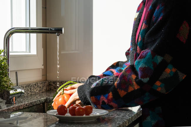 Обрізане зображення жінки в барвистій куртці, що миє свіжий помідор під чистою водою над раковиною на кухні вдома — стокове фото