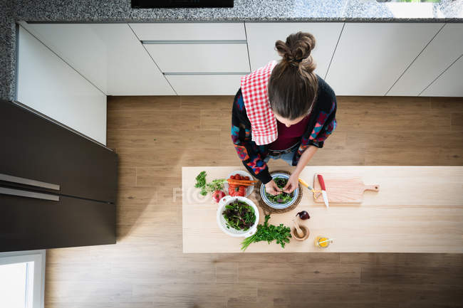 Femme en veste multicolore préparant des légumes tout en cuisinant une salade saine dans la cuisine — Photo de stock