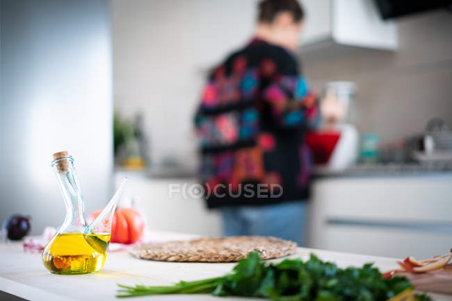 Olive oil in modern jar with anonymous lady in multicolored jacket preparing healthy salad in kitchen — Stock Photo