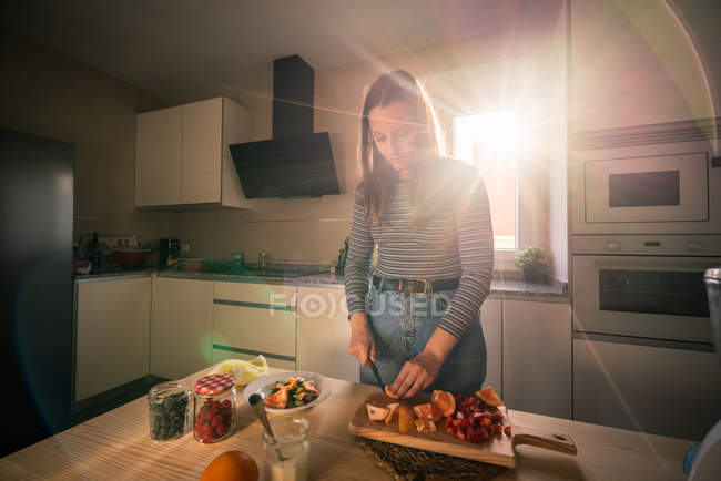 Junge Frau in lässigem Outfit schneidet frisches Obst, während sie in der gemütlichen Küche unter Sonnenstrahlen kocht — Stockfoto