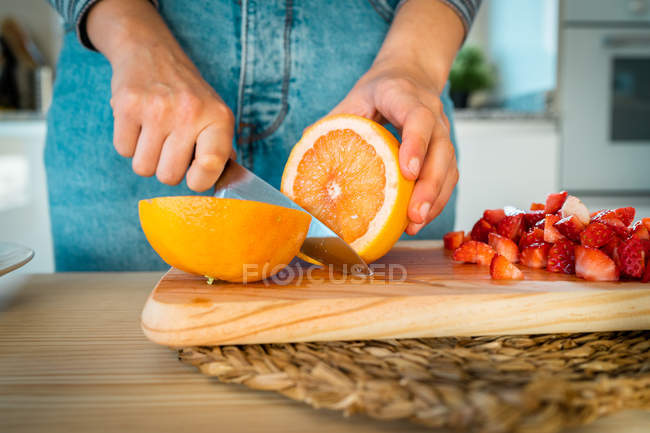 Imagen recortada de la mujer en traje casual cortar naranja fresca cerca de trozos de fresa cortada - foto de stock