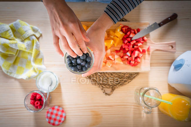 Imagem cortada de mulher tomando mirtilos do frasco enquanto cozinha alimentos saudáveis de vitaminas de frutas frescas em casa — Fotografia de Stock