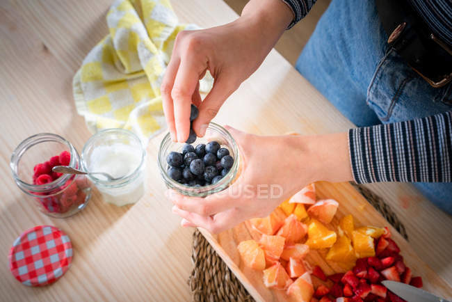 Geschnittenes Bild einer Frau, die Blaubeeren aus dem Glas nimmt, während sie zu Hause gesunde Vitaminnahrung aus frischen Früchten kocht — Stockfoto