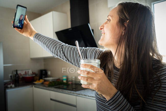 Jovem com jarra de iogurte saudável sorrindo e posando para selfie na cozinha — Fotografia de Stock