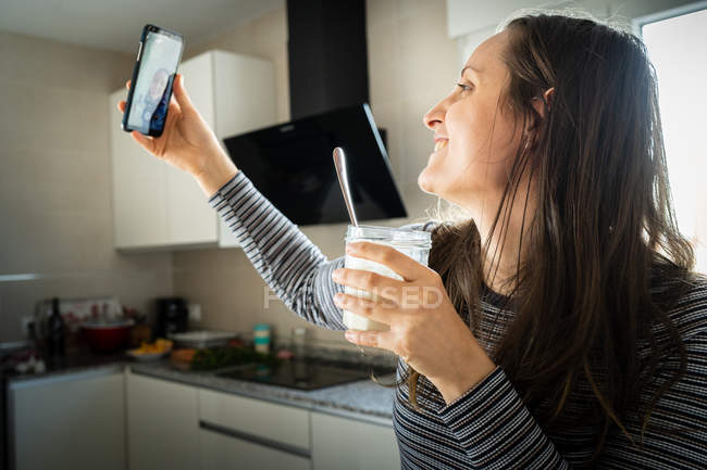 Junge Frau mit Glas mit gesundem Joghurt lächelt und posiert für Selfie in Küche — Stockfoto