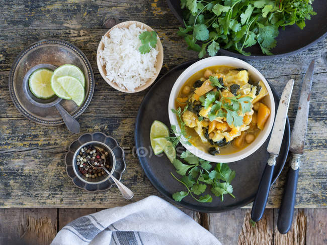 Plato con deliciosos curry vegetariano y rodajas de lima colocadas en bandeja sobre fondo de madera junto a arroz, pimienta y limas frescas - foto de stock