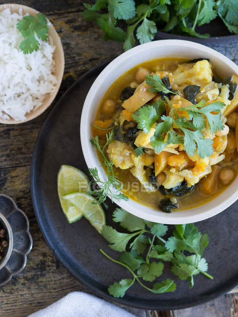 Assiette avec délicieux curry végétarien et tranches de citron vert placés sur un plateau sur fond en bois à côté de riz et de citron vert frais — Photo de stock