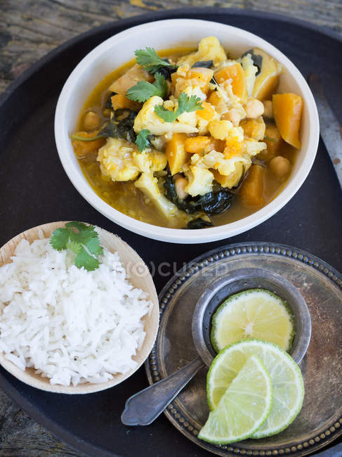 Assiette avec délicieux curry végétarien et tranches de citron vert placés sur un plateau sur fond en bois à côté de riz et de limes fraîches — Photo de stock