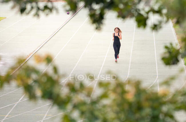 Vista entre los árboles de una joven en forma de mujer corriendo en una pista al aire libre en un día soleado - foto de stock