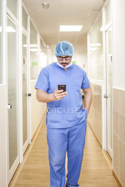 Чоловічий хірург стоїть в коридорі під час перевірки повідомлень на своєму смартфоні — стокове фото