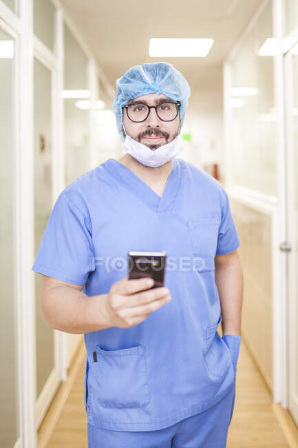 Cirujano masculino de pie en el pasillo mientras revisa los mensajes en su teléfono inteligente, mira la cámara - foto de stock