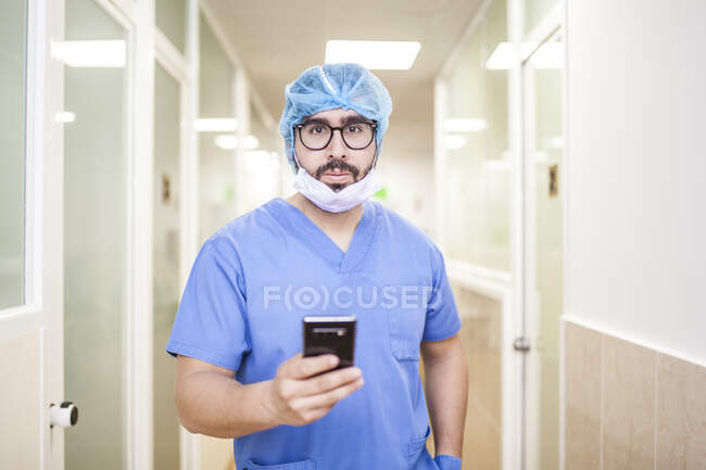 Cirujano masculino de pie en el pasillo mientras revisa los mensajes en su teléfono inteligente, mira la cámara - foto de stock