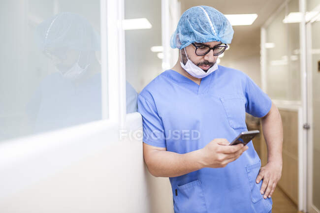 Cirujano masculino apoyado en la pared del pasillo mientras comprueba los mensajes en su teléfono inteligente - foto de stock