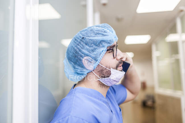 Cirujano masculino apoyado en la pared del pasillo mientras habla con su teléfono inteligente, vista lateral - foto de stock