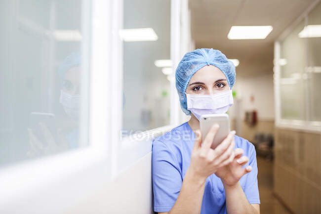 Chirurgin steht auf dem Flur, während sie Nachrichten auf ihrem Smartphone checkt, blickt in die Kamera und lächelt — Stockfoto