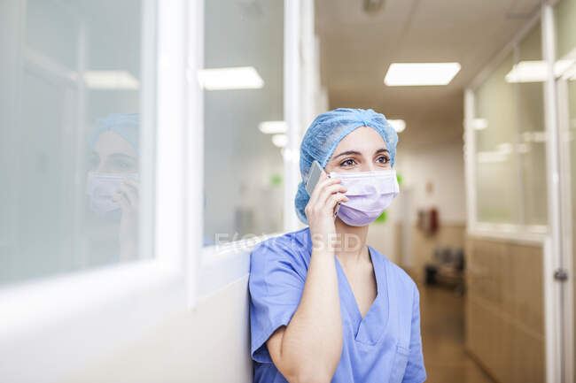 Жінка-хірург спирається на коридорну стіну, розмовляючи зі своїм смартфоном — стокове фото