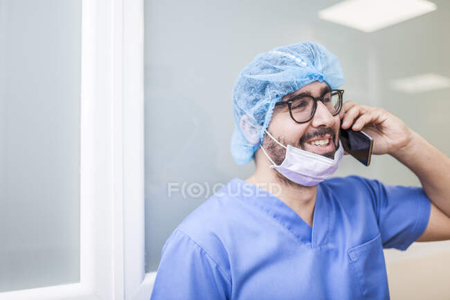 Cirurgião do sexo masculino inclinado na parede do corredor enquanto conversa com seu telefone inteligente — Fotografia de Stock