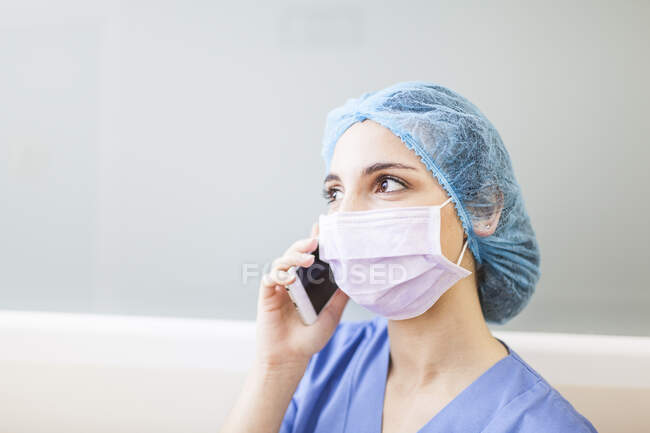 Жінка-хірург спирається на коридорну стіну, розмовляючи зі своїм смартфоном — стокове фото