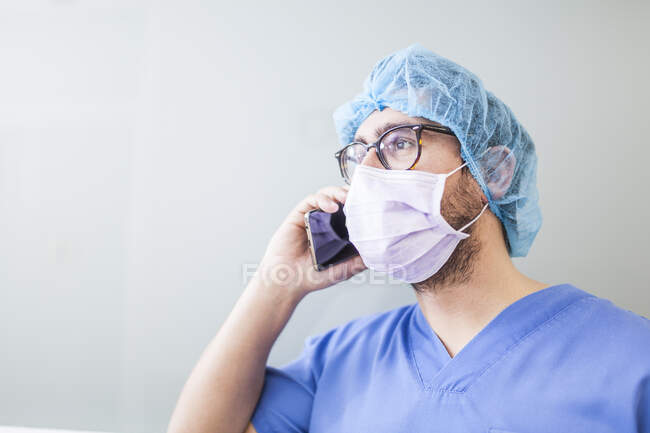 Chirurgo maschio appoggiato sulla parete del corridoio mentre parla con il suo smartphone — Foto stock