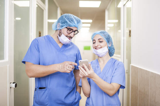 Колеги хірурги чоловік і жінка спілкуються під час ходьби до операційної, вона показує йому свій смартфон і посміхається — стокове фото