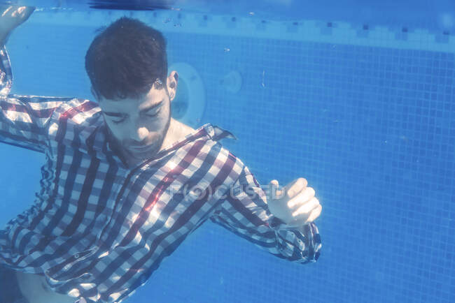 Hombre con camisa posando con los ojos cerrados bajo el agua en la piscina. - foto de stock
