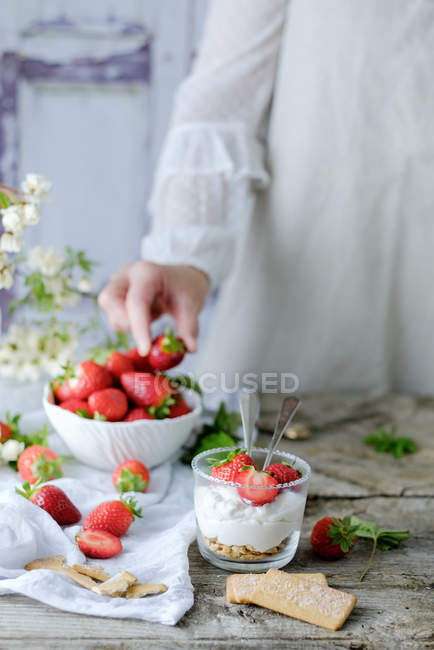 Сливочный сладкий десерт со свежей сочной клубникой, подаваемый в стакане на деревенском деревянном столе с печеньем и женской рукой, берущей ягоды на заднем плане — стоковое фото