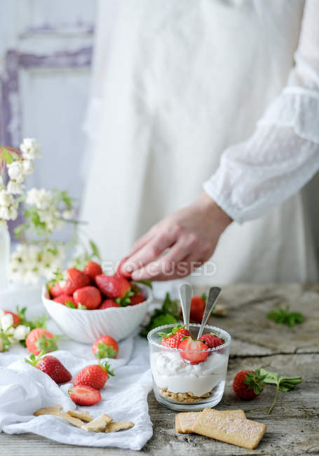 Creamy sweet dessert with fresh juicy strawberries served in glass on rustic wooden table with biscuits and female hand taking berries on background — Stock Photo