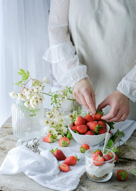 Creamy sweet dessert with fresh juicy strawberries served in glass on rustic wooden table with biscuits and female hand taking berries on background — Stock Photo