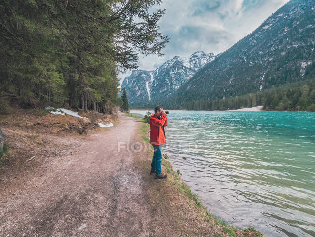 Seitenansicht eines erwachsenen Reisenden, der Fotos von Bergrücken macht, während er am Ufer des ruhigen Sees in der Natur steht — Stockfoto