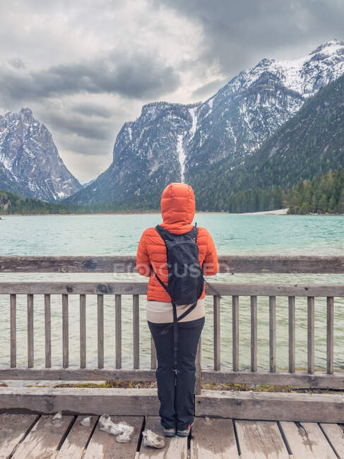Unrecognizable tourist in warm clothes standing on wooden pier and looking at mountain ridge and rippling lake water on cloudy day in countryside — Stock Photo