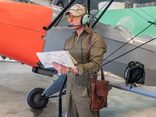 Piloto sosteniendo mapa cerca de avión pequeño en hangar - foto de stock