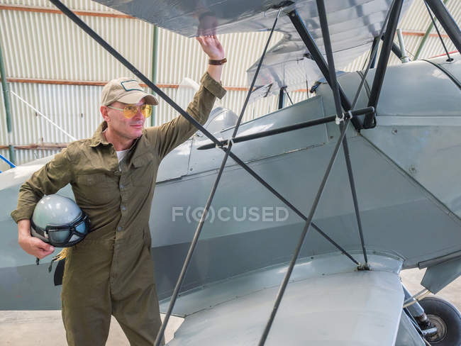 Confident pilot standing near retro plane in hangar and holding helmet — Stock Photo