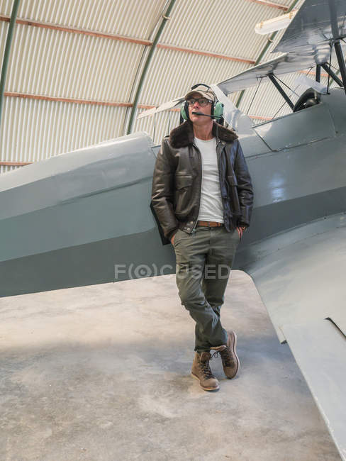 Confident pilot with headset leaning on retro plane in hangar — Stock Photo