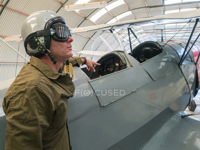 Piloto masculino em equipamento de pé ao lado de biplano vintage em hangar — Fotografia de Stock