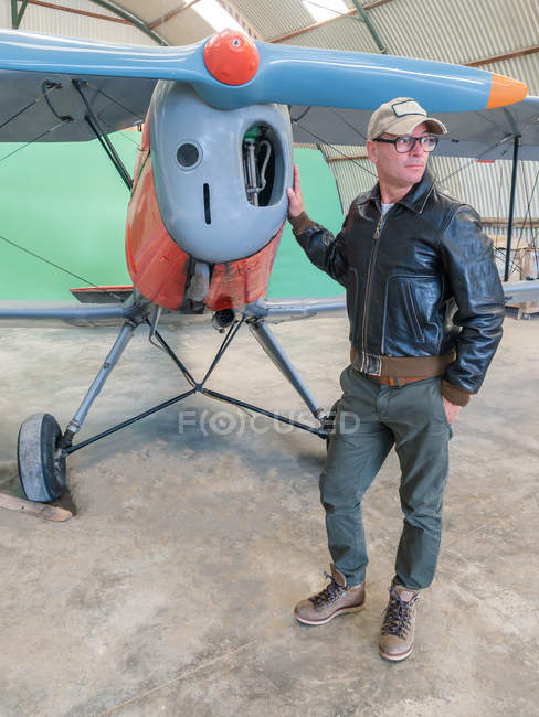Confident pilot standing next to retro plane in hangar and looking away — Stock Photo