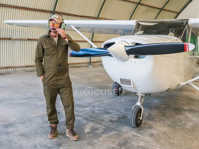 Уверенный пилот в гарнитуре, стоящий рядом с ретро-самолетом в ангаре — стоковое фото