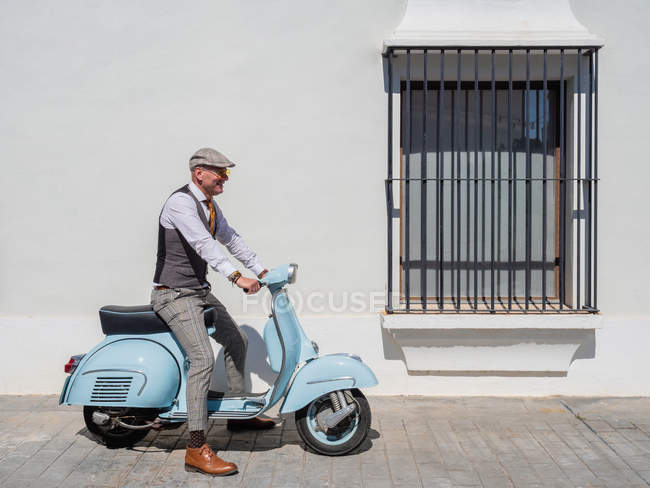 Hipster positivo de mediana edad en ropa elegante con moto retro mirando hacia otro lado en un día soleado - foto de stock