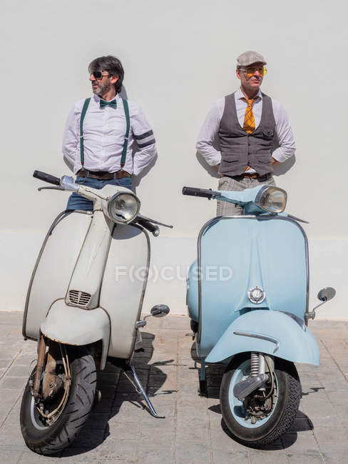 Dreamy middle-aged hipsters in fashionable clothes with retro motorbikes leaning on wall in sunny day — Stock Photo