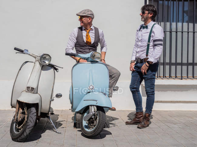 Positive middle-aged hipsters in elegant clothes with retro motorbikes looking away in sunny day — Stock Photo