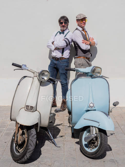 Dreamy middle-aged hipsters in fashionable clothes with retro motorbikes leaning on wall posing in sunny day — Stock Photo