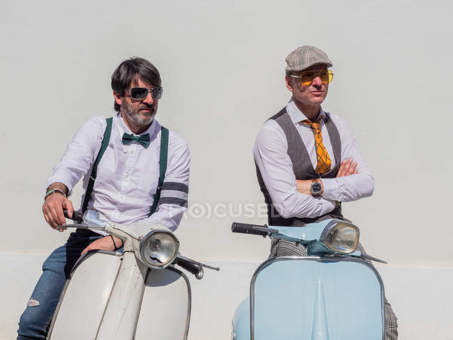 Hipsters positivos de mediana edad en ropa elegante con motos retro mirando hacia otro lado en un día soleado - foto de stock